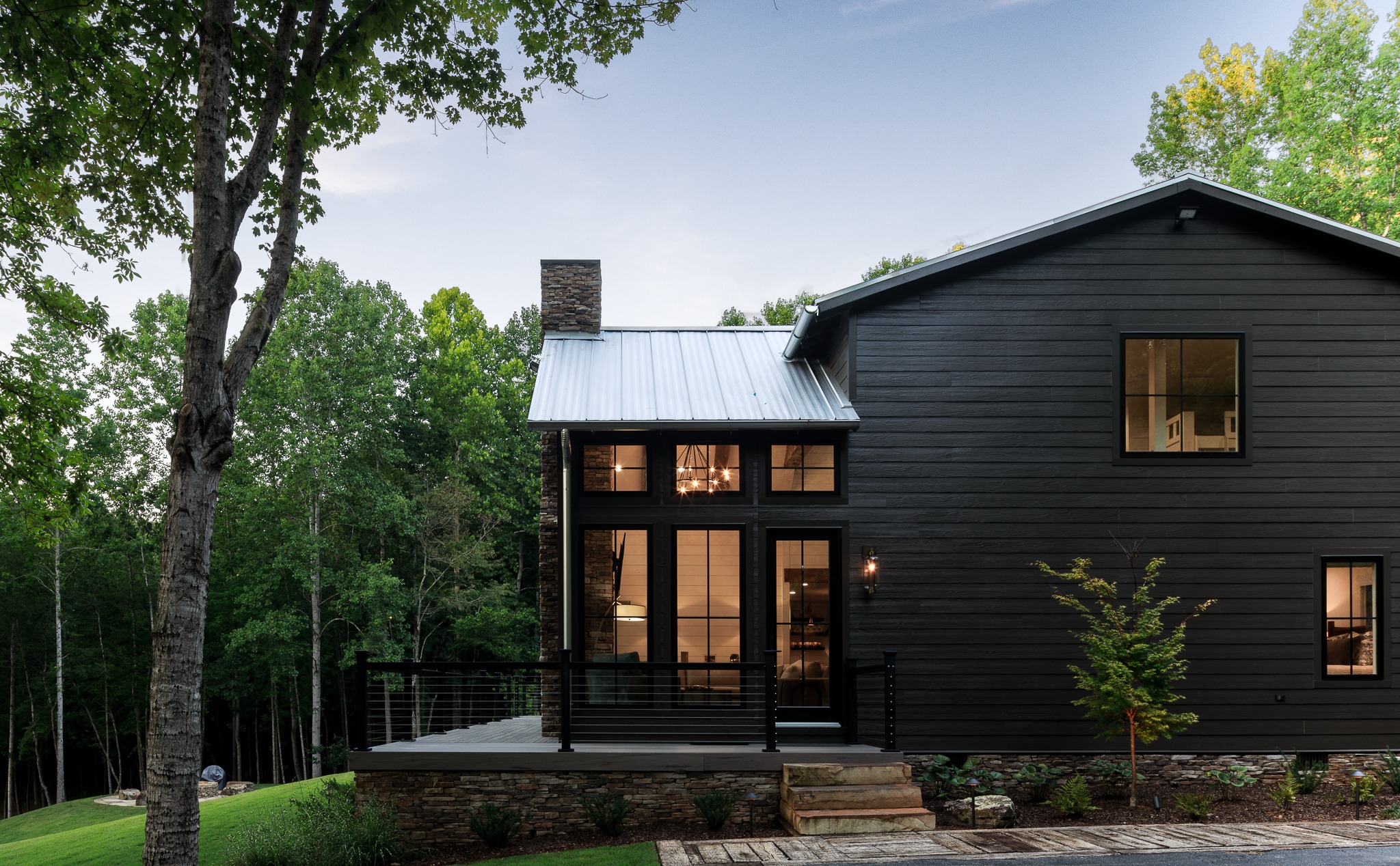 Dark modern farmhouse at dusk, windows glowing through the trees